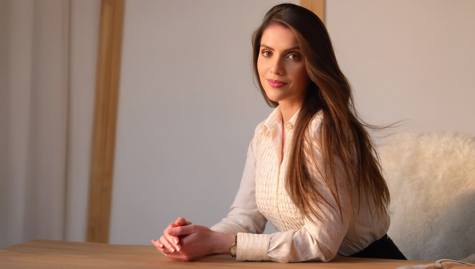 Mulher adulta em ambiente interno, sentada à mesa de madeira clara. Ela apoia os antebraços sobre a mesa, com as mãos juntas à frente do corpo, e olha diretamente para quem observa, com expressão calma e confiante. Tem pele clara, cabelos longos e lisos em tom castanho, repartidos de lado e caindo sobre os ombros. Veste uma blusa clara de mangas longas, com textura suave e discreta. Está sentada em uma cadeira estofada clara, com aparência aconchegante. O fundo é simples e desfocado, em tons neutros de bege e madeira, com iluminação suave e quente, criando um clima acolhedor e sereno.