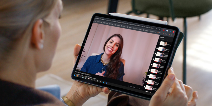 Pessoa vista de trás e de lado segura um tablet com as duas mãos, em um ambiente interno com piso de madeira claro e cadeiras desfocadas ao fundo. Na tela do tablet, aparece uma mulher adulta em uma videochamada ou gravação: ela está centralizada, sorrindo, com expressão acolhedora, pele clara e cabelos longos e castanhos, soltos sobre os ombros. Veste uma blusa azul de mangas longas. O enquadramento da tela mostra um fundo neutro e claro atrás dela. À direita da tela do tablet, há uma coluna vertical com várias miniaturas pequenas, alinhadas uma abaixo da outra, indicando outros vídeos ou cenas. Na parte superior da tela, há uma barra escura com elementos de interface e textos pequenos, não legíveis. A iluminação geral é suave e natural, transmitindo um clima calmo e profissional.