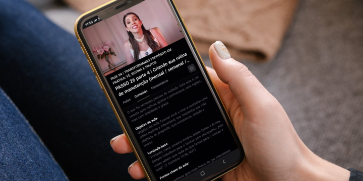 Pessoa segura um smartphone na vertical com a mão direita, enquanto está sentada, com parte das pernas cobertas por tecido escuro visível ao fundo. Na tela do celular, há um site em fundo preto. No topo, aparece uma imagem pequena de uma mulher adulta sorrindo, com cabelos longos e castanhos, vestindo roupa clara, em um ambiente interno. Abaixo da imagem, há vários blocos de texto em letras brancas e cinza, organizados em parágrafos e listas, com títulos e subtítulos em destaque; o conteúdo textual é extenso, mas não está legível em detalhe. A interface sugere leitura de um artigo, conteúdo educativo ou informativo. O ambiente ao redor é aconchegante, com um tapete claro desfocado ao fundo, e a iluminação é suave e natural.